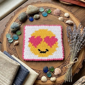 May include: A square, white crocheted coaster with a yellow emoji face featuring pink heart eyes and a black smile. The coaster has a pink border. The image also includes decorative elements like stones, shells, and fabric.