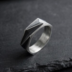 Könnte beinhalten: Ein silberfarbener Ring mit einer geometrischen, facettierten Oberfläche und einem dunklen, gebürsteten Finish. Die Ringschiene hat eine glatte, polierte Innenseite und ein strukturiertes, kantiges Außendesign mit scharfen Flächen.