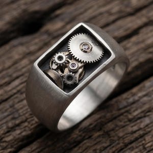 May include: A silver-toned ring featuring a rectangular face with an open setting that displays a cluster of small, intricate metallic gears and cogs. The band has a brushed finish, and the internal mechanism components are silver and bronze.