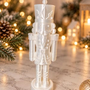 Könnte beinhalten: Eine weiße Nussknacker-Figur mit glänzender Oberfläche, die einen hohen Hut, eine Uniform mit Knöpfen und einen Bart aufweist. Der Nussknacker steht auf einem runden Sockel und zeigt detaillierte, geformte Verzierungen an der Jacke.