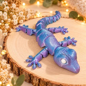Puede incluir: Una figura de lagarto articulada impresa en 3D con un acabado iridiscente brillante que cambia entre tonos azules y morados. El lagarto tiene un cuerpo segmentado, una cola larga, cuatro patas detalladas con dedos extendidos y grandes ojos blancos.
