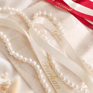 May include: A collection of cream-colored satin ribbons featuring gold-printed names Aylin and Furkan, alongside red satin ribbons with gold-printed names Dclal and Ramil, intertwined with a long strand of white faux pearls on a textured surface.