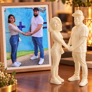Può includere: Una fotografia incorniciata di una coppia che si tiene per mano davanti a un muro blu, esposta accanto a una statuetta coordinata stampata in 3D color bianco sporco della stessa coppia in posa identica, su una superficie in legno.