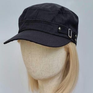 May include: A black military-style cadet cap featuring a flat top, a curved brim, and a decorative side strap with a silver-toned metal buckle and two small silver-toned rivets. The cap is displayed on a mannequin head with blonde hair.