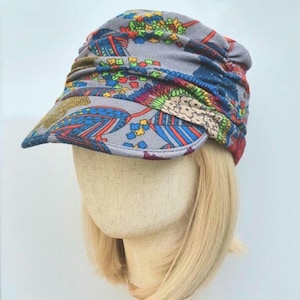 May include: A grey newsboy cap featuring a vibrant, multi-coloured floral and geometric pattern. The hat has a gathered, ruched crown design and a stiff, curved brim. The fabric displays bright shades of blue, red, green, and yellow throughout.