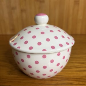 May include: A white ceramic sugar bowl with a lid, decorated with pink polka dots. The lid features a small, round handle, white with a pink top. The bowl is on a wooden surface.