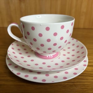 Puede incluir: Juego de taza y platillo blancos con lunares rosas. La taza tiene un asa curva y se asienta sobre un platillo a juego y un plato más pequeño. El juego es de cerámica y está diseñado para servir té.