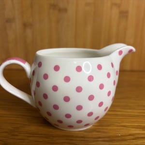 以下が含まれることがあります： ピンクのドット模様が施された白いセラミック製のクリーマー。クリーマーは、曲がったハンドルと注ぎ口を備えています。ハンドルと注ぎ口の縁もピンク色です。クリーマーは木製の表面に置かれています。