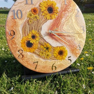 Może przedstawiać: Okrągły zegar ścienny ze złotym tłem z żywicy i osadzonymi słonecznikami. Srebrne cyfry oznaczają godziny. Wskazówki zegara są srebrne. Zegar jest wystawiony na zewnątrz na trawiastym terenie.