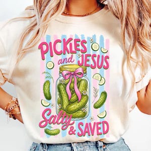 May include: Cream-colored t-shirt with a graphic of a jar of pickles tied with a pink bow, surrounded by pickle slices and flowers. The text "PICKLES and JESUS Salty & SAVED" is displayed in pink and green.