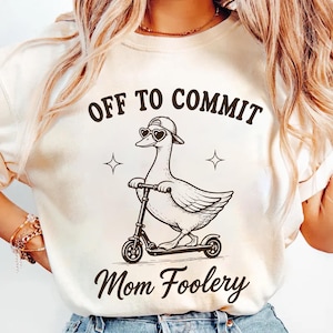 Può includere: Felpa color crema con la scritta "OFF TO COMMIT Mom Foolery". La grafica presenta un'anatra con occhiali da sole a forma di cuore e un berretto, su uno scooter. Il disegno è in inchiostro nero.