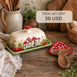 May include: A ceramic butter dish with a lid and a matching mushroom-shaped spoon. The dish is off-white with a green base and features a hand-painted design of red mushrooms and foliage. The lid has a red mushroom knob. The spoon is red with white spots.