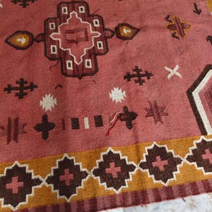 Pu&ograve; includere: Un grande tappeto rettangolare con sfondo rosso sbiadito e motivi geometrici marroni, bianchi e gialli. Il tappeto presenta un disegno centrale e un bordo a motivi, che mostra un'estetica tradizionale.
