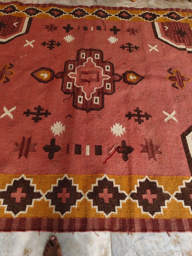 Pu&ograve; includere: Un grande tappeto rettangolare, di colore rosa-rossastro con motivi geometrici. Il design comprende forme marroni, bianche e gialle. Il bordo presenta un motivo a rombi ripetuto. Il tappeto presenta croci, stelle e altre forme.