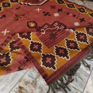 Pu&ograve; includere: Un tappeto rettangolare fatto a mano con un motivo geometrico nei toni del rosso, marrone, giallo e bianco. Il tappeto presenta un disegno centrale con forme ripetute e un bordo con frange. Il tappeto &egrave; realizzato in un materiale morbido e tessuto.