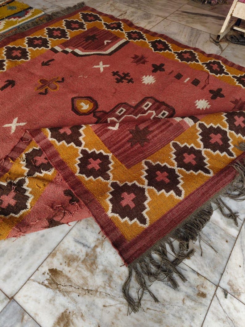 Pu&ograve; includere: Un tappeto rettangolare fatto a mano con un motivo geometrico nei toni del rosso, marrone, giallo e bianco. Il tappeto presenta un disegno centrale con forme ripetute e un bordo con frange. Il tappeto &egrave; realizzato in un materiale morbido e tessuto.