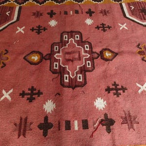 Pu&ograve; includere: Un tappeto tessuto a mano con un design geometrico centrale nei toni del rosso, marrone e bianco. Il tappeto presenta un bordo con motivi ripetuti in marrone e giallo, adatto per l'arredamento della casa.