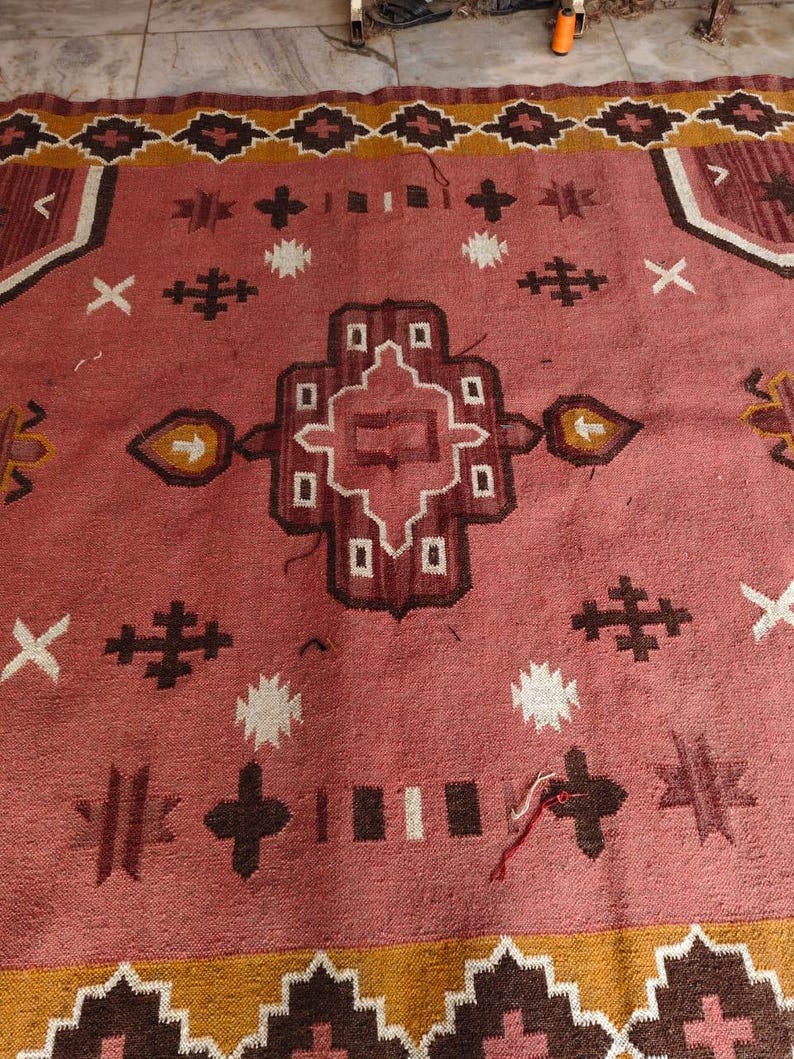Pu&ograve; includere: Un tappeto tessuto a mano con un design geometrico centrale nei toni del rosso, marrone e bianco. Il tappeto presenta un bordo con motivi ripetuti in marrone e giallo, adatto per l'arredamento della casa.
