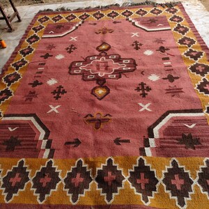 Pu&ograve; includere: Un grande tappeto rettangolare con un motivo geometrico centrale nei toni del rosso, marrone e bianco. Il tappeto presenta un bordo di forme a diamante ripetute in marrone e oro, con bordi sfrangiati.