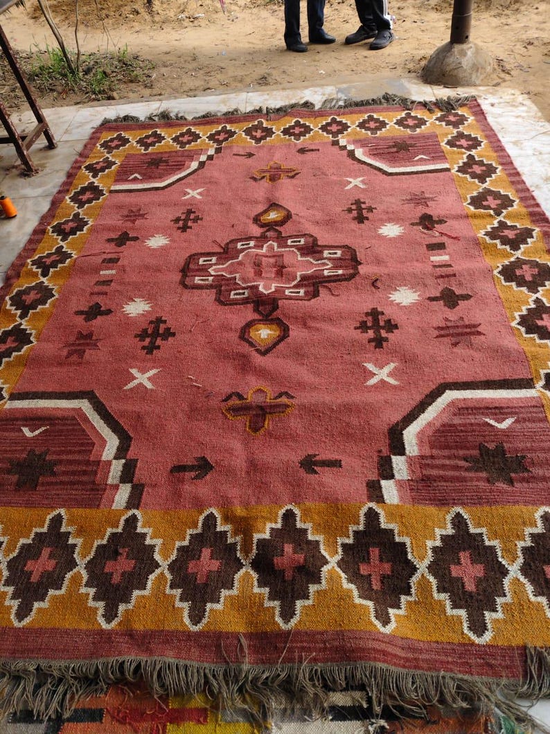 Pu&ograve; includere: Un grande tappeto rettangolare con un motivo geometrico centrale nei toni del rosso, marrone e bianco. Il tappeto presenta un bordo di forme a diamante ripetute in marrone e oro, con bordi sfrangiati.