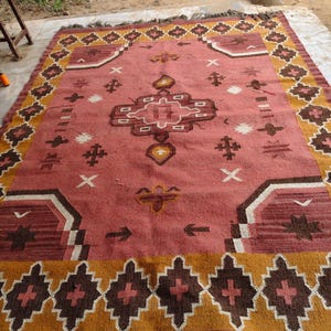 Pu&ograve; includere: Un tappeto rettangolare con un design geometrico centrale nei toni del rosso, marrone e bianco. Il tappeto presenta un bordo di forme a diamante marrone e oro e frange lungo i bordi. Il tappeto &egrave; posto su una superficie di cemento.