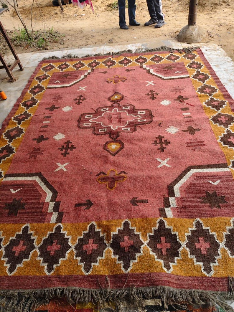 Pu&ograve; includere: Un tappeto rettangolare con un design geometrico centrale nei toni del rosso, marrone e bianco. Il tappeto presenta un bordo di forme a diamante marrone e oro e frange lungo i bordi. Il tappeto &egrave; posto su una superficie di cemento.