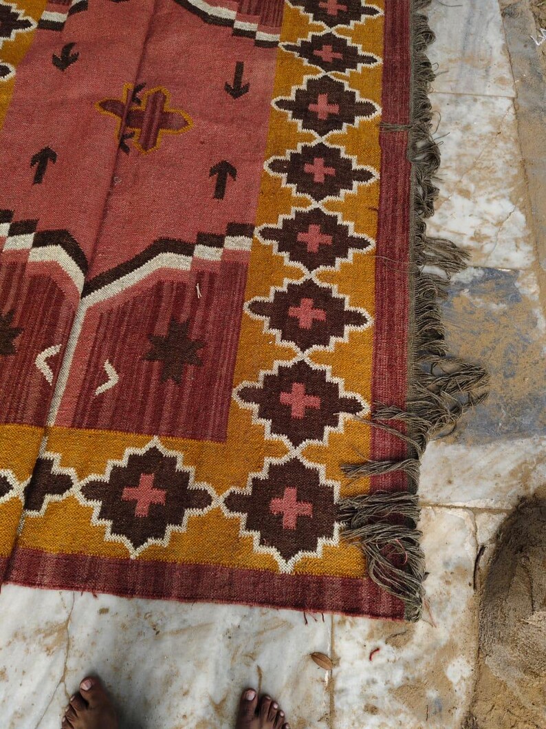 Pu&ograve; includere: Un tappeto rettangolare con un motivo geometrico nei toni del rosso, marrone, giallo e bianco. Il tappeto presenta un bordo con forme a diamante e frange lungo un bordo. Il design include frecce e motivi a forma di croce.