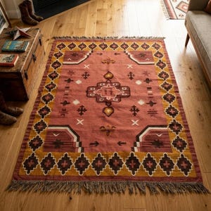 Pu&ograve; includere: Un tappeto rettangolare in stile sud-ovest, con un campo centrale color terracotta. Presenta motivi geometrici marroni, dorati e bianchi e ha un bordo sfrangiato. Il tappeto &egrave; posato su un pavimento in legno.