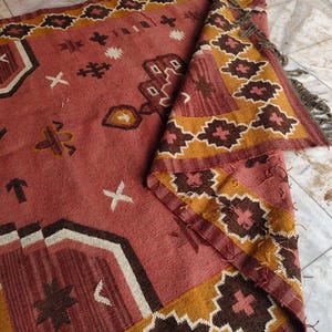 Pu&ograve; includere: Un tappeto rettangolare piegato con un motivo geometrico. Il tappeto presenta un campo centrale rosa-rossastro con forme geometriche marroni, bianche e gialle. Il bordo presenta un motivo ripetuto di disegni floreali marroni e gialli. Le frange sono visibili sul bordo.