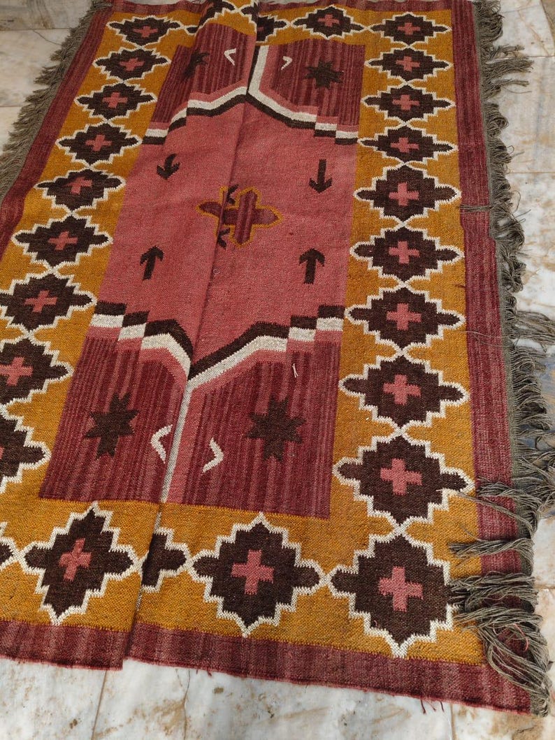 Pu&ograve; includere: Un tappeto rettangolare con un motivo geometrico nei toni del rosso, marrone e oro. Il tappeto presenta un campo centrale rosso con un disegno geometrico e un bordo di forme a diamante ripetute. I bordi hanno una frangia.