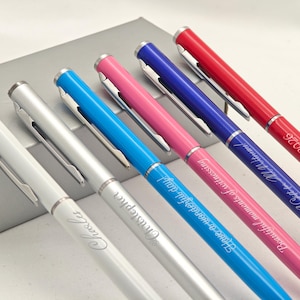 May include: Six coloured pens with silver clips and tips. The pens are white, silver, blue, pink, purple, and red. Some pens are engraved with names and phrases, including "Charles," "Christopher," and "June 6, 2026."