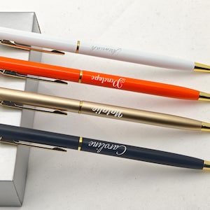 Op de afbeelding: Vier gepersonaliseerde pennen in wit, oranje, goud en marineblauw, elk met gouden accenten. De pennen zijn gegraveerd met namen in een cursief lettertype. De pennen zijn gerangschikt op een lichtgrijze ondergrond.