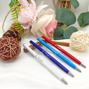 May include: Four personalized pens in white, blue, light blue, and red, each with a silver tip. The pens are engraved with names in a cursive font. The pens are arranged on a white surface with decorative floral and natural elements.