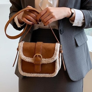 May include: A brown suede crossbody bag with a shearling trim and a gold-tone buckle. The bag has a long, adjustable strap and a flap closure. The bag is being held by a person wearing a gray blazer and a white blouse.