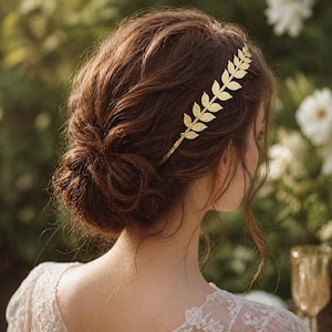 May include: A gold-colored leaf hair comb is worn in the hair. The hair is styled in a low bun. The comb is made of metal and has a series of detailed leaves. The background is blurred, with greenery and white flowers.