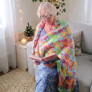 Könnte beinhalten: Eine grob gestrickte Überwurfdecke mit einem lebendigen, mehrfarbigen Garnmuster in Pastelltönen wie Rosa, Orange, Gelb, Grün und Violett. Die Decke hat eine weiche, strukturierte und flauschige Optik mit einer dicken, geschlungenen Strickkonstruktion.