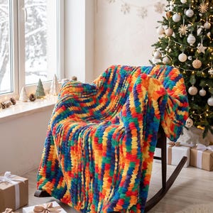 Pode incluir: Uma manta de malha grossa com um padrão vibrante de riscas multicoloridas em tons de vermelho, laranja, amarelo, verde e azul. O fio macio e texturizado cria uma aparência felpuda e em laços, drapeada sobre uma cadeira de balanço.