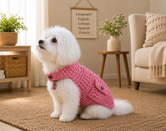 May include: A small white dog with fluffy fur wearing a bright pink crocheted sweater. The sweater features a textured knit pattern, a high collar, and a small pocket on the side. A dark button is visible on the collar and on the pocket flap.