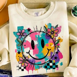 Könnte beinhalten: Cremefarbenes Sweatshirt mit einem großen, farbenfrohen Smiley-Gesicht-Design. Die Grafik umfasst einen schwarzen Umriss, Blumen, Sterne und ein Schachbrettmuster. Das Smiley-Gesicht ist mit leuchtenden Farben wie Pink, Blau, Gelb und Lila gefüllt.