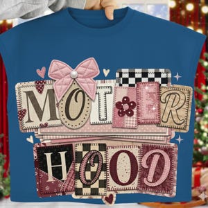 Op de afbeelding: Een blauw t-shirt met het woord "MOTHERHOOD" in een patchwork-ontwerp. De letters zijn in verschillende kleuren en patronen, waaronder roze, bordeauxrood en zwart-wit geblokt. Een roze strik en hartjes versieren het shirt.