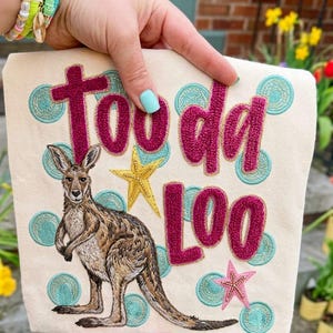 May include: Beige canvas tote bag with the embroidered words "Too da Loo" in magenta. A kangaroo and starfish are also embroidered on the bag. Teal and gold accents surround the text and images. The bag is held by a person wearing colorful bracelets.