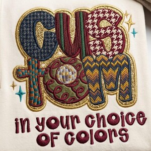 Könnte beinhalten: Cremefarbenes Sweatshirt mit einem gestickten Design. Das Design zeigt das Wort "CUSTOM" in verschiedenen Mustern und Farben, darunter der Text "in your choice of colors". Goldglitzer umrandet die Buchstaben.
