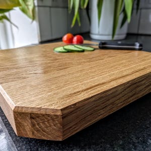 May include: An octagonal wooden chopping board with a natural wood grain. Two red cherry tomatoes and three cucumber slices are on the board, along with a black knife. The board is on a dark countertop.