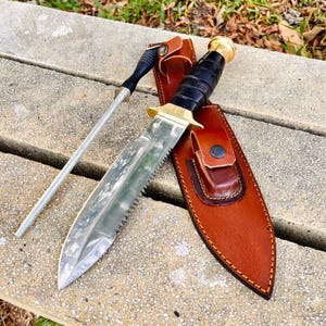 May include: A survival knife with a serrated blade, black handle, and brass detailing, presented in a brown leather sheath. A sharpening steel is also visible. The knife and sheath are on a concrete surface.
