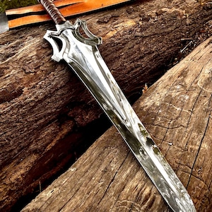 Puede incluir: Una espada pulida de color plateado con un mango envuelto en cuero marrón y una funda a juego. La hoja tiene una guarda decorativa y descansa sobre una superficie de madera envejecida. El diseño de la espada sugiere un tema de fantasía o histórico.