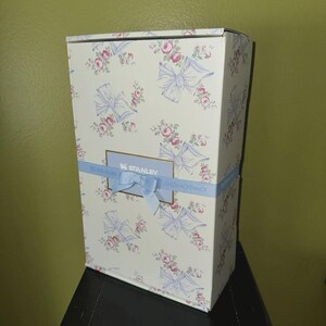May include: A rectangular gift box featuring a floral design of pink roses and pale blue bows on a white backdrop. A pale blue ribbon with a bow encircles the box, with the word "STANLEY" visible. The box is on a black surface.