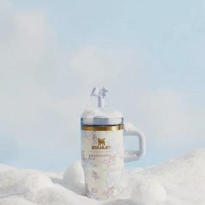 May include: A white Stanley travel mug with a floral design and a handle. The mug has a gold rim and a clear lid with a decorative topper. The mug is set in a snowy scene.