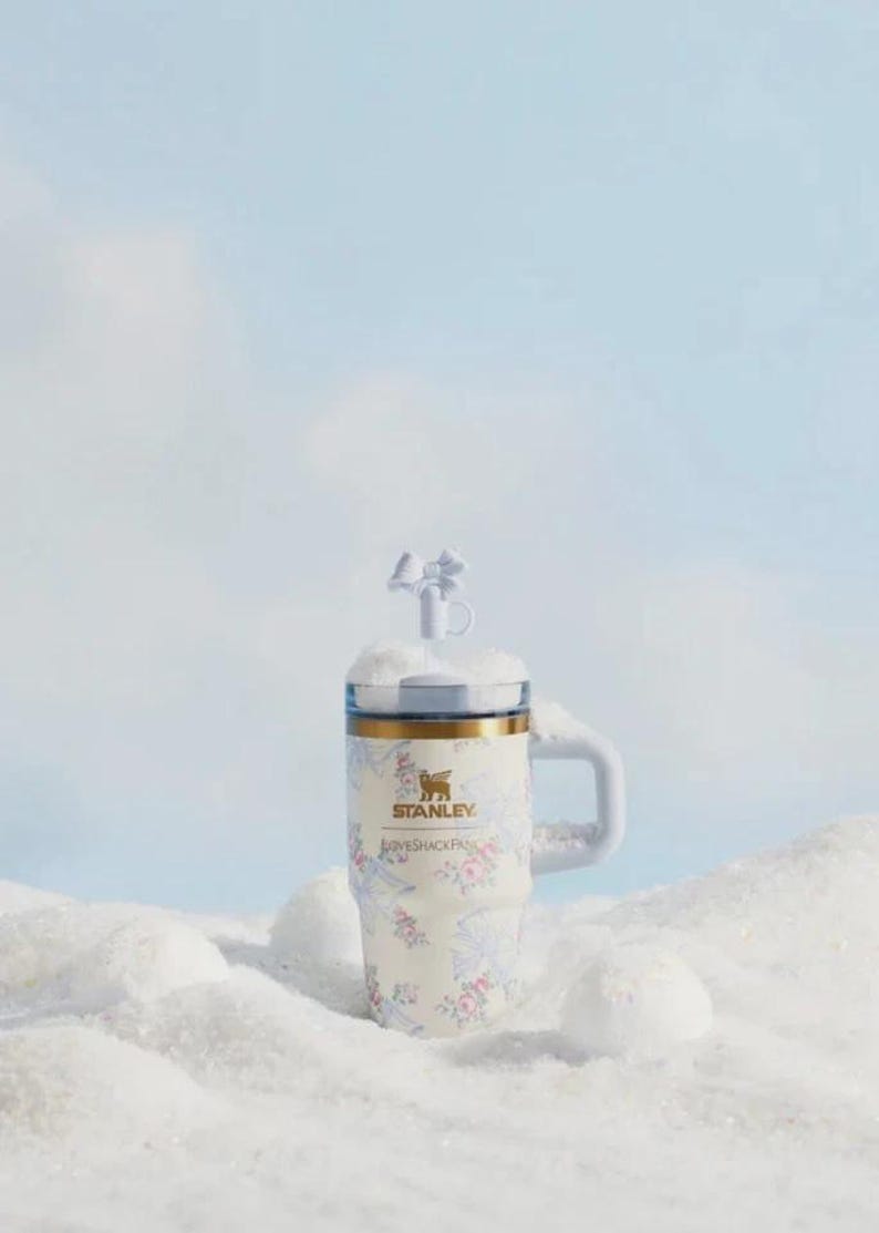 May include: A white Stanley travel mug with a floral design and a handle. The mug has a gold rim and a clear lid with a decorative topper. The mug is set in a snowy scene.