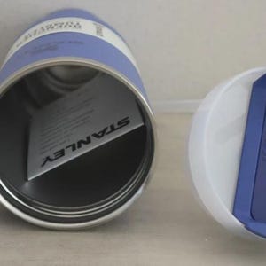 Puede incluir: Un termo Stanley azul y blanco con asa y tapa blanca con detalle azul y pajita. El termo tiene interior plateado y una etiqueta en el interior con el logo Stanley.