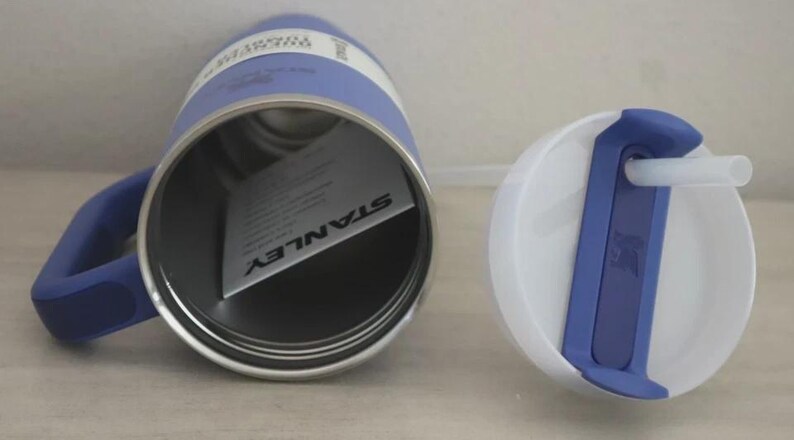 Puede incluir: Un termo Stanley azul y blanco con asa y tapa blanca con detalle azul y pajita. El termo tiene interior plateado y una etiqueta en el interior con el logo Stanley.